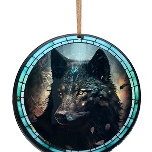 May include: Round ornament featuring a detailed illustration of a black wolf with yellow eyes. The wolf is set against a dark, starry background within a teal and black stained-glass style border. A gold ribbon is attached for hanging.