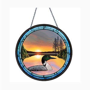May include: A round stained glass window hanging from a chain. The window depicts a loon swimming in a lake at sunset. The lake is surrounded by trees and the sky is a mix of orange, pink, and blue.