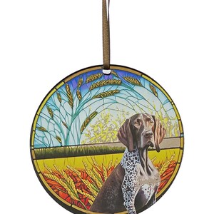 May include: Round ornament featuring a German Shorthaired Pointer dog in a field of wheat. The dog is brown and white with a focused expression. The background has a stained-glass effect with blue, yellow, and green hues. A brown ribbon is attached for hanging.