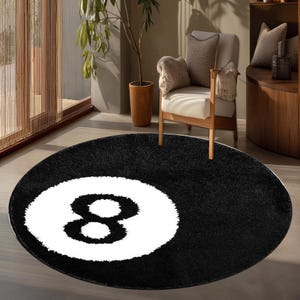 May include: A circular area rug featuring a black background with a large, centered white circle containing a black number eight. The rug has a soft, textured pile surface and is designed in the likeness of a classic eight ball from a billiards set.