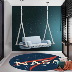 Puede incluir: Un sofá colgante de color azul claro suspendido por correas blancas desde el techo. Debajo del sofá hay una alfombra redonda de color azul oscuro con el logotipo de la NASA en blanco y rojo, una órbita estilizada y estrellas blancas.