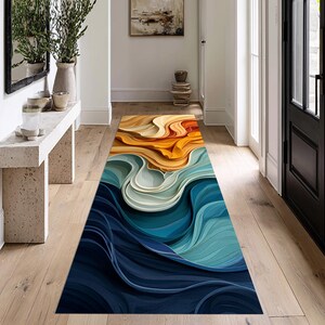 May include: A rectangular rug with an abstract design. The rug has a flowing pattern of layered colours, transitioning from deep navy blue at the bottom to shades of teal, white, orange, and yellow at the top. The rug is placed on a wooden floor.