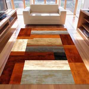 Pode incluir: Um tapete retangular com um design geométrico. O tapete apresenta um padrão de formas retangulares em tons de laranja, castanho, bege, cinza e preto. Está colocado sobre um chão de madeira, com um sofá branco ao fundo.