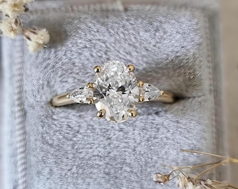 May include: A gold ring featuring a large oval-cut center stone flanked by two smaller pear-cut stones. The ring is set into a grey velvet ring box, with small dried white flowers positioned near the corners of the square-shaped box.