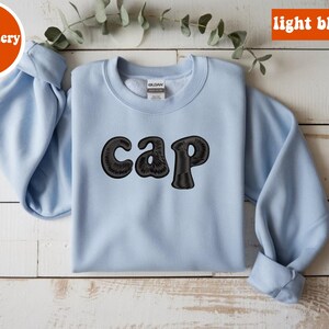 May include: A light blue sweatshirt with the word "cap" embroidered in black. The word "embroidery" is in a circle in the upper left corner, and "light blue" is in a rectangle in the upper right corner.