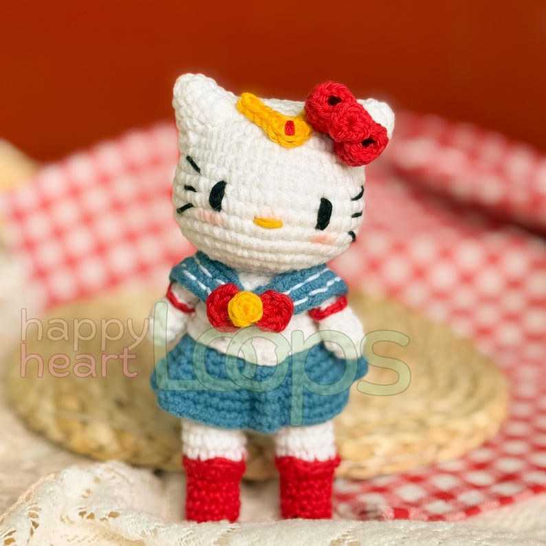 Może przedstawiać: Biała, szydełkowa lalka Hello Kitty w niebieskim stroju marynarskim z czerwoną kokardką i ż&oacute;łtą gwiazdą na głowie.