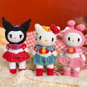 Puede incluir: Tres muñecas de ganchillo hechas a mano. Las muñecas tienen forma de personajes de dibujos animados. Una muñeca es negra con orejas puntiagudas, otra es blanca con un lazo rojo y la tercera es rosa con una capucha. Las muñecas visten trajes con detalles rojos, azules y blancos. El texto "happy heart" es visible.