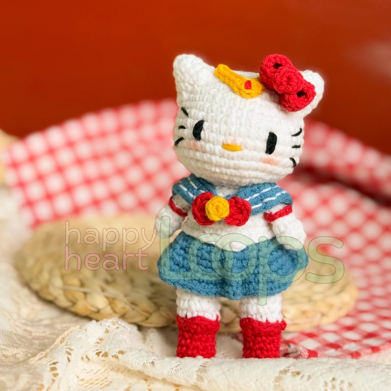 Może przedstawiać: Biała pluszowa lalka Hello Kitty w niebieskiej sukience marynarskiej z czerwoną kokardką i ż&oacute;łtym kwiatkiem. Lalka stoi na białym, dzianym podkładzie.
