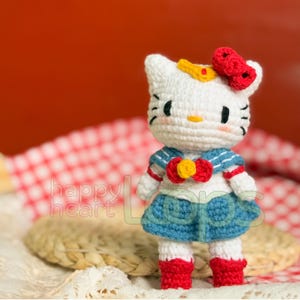 Może przedstawiać: Biała, szydełkowa lalka Hello Kitty w niebieskiej marynarskiej sukience z czerwoną kokardką i ż&oacute;łtą gwiazdą. Lalka ma czerwoną kokardkę na głowie i stoi na wiklinowym koszu.