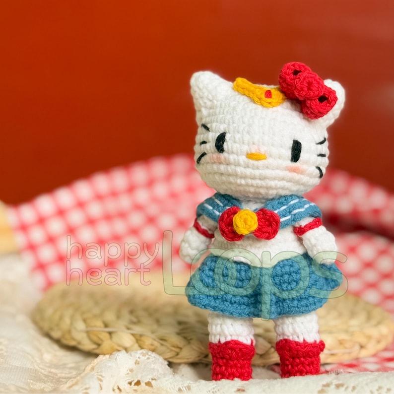 Może przedstawiać: Biała, szydełkowa lalka Hello Kitty w niebieskiej marynarskiej sukience z czerwoną kokardką i ż&oacute;łtą gwiazdą. Lalka ma czerwoną kokardkę na głowie i stoi na wiklinowym koszu.