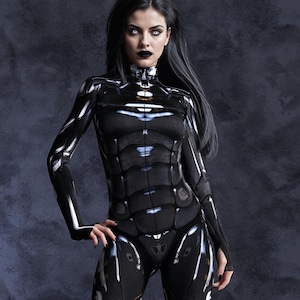 Puede incluir: Una mujer con un traje futurista negro y plateado de diseño elegante y ajustado. El traje tiene un acabado texturizado y metálico y presenta un cuello alto y mangas largas.