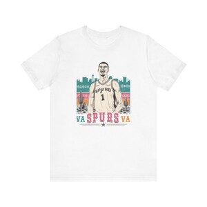May include: White t-shirt featuring a graphic of a basketball player in a Spurs jersey with the number 1. The design includes a desert landscape with cacti and the text "VA SPURS VA".
