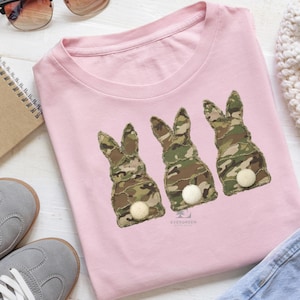 Puede incluir: Camiseta rosa claro con tres siluetas de conejos hechas de tela de camuflaje y colas de pompón blancas. La camiseta tiene cuello redondo y está hecha de un material suave. El diseño está centrado.