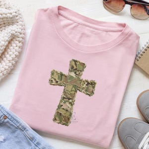May include: A light pink t-shirt featuring a camouflage cross applique. The cross is made of various shades of green, brown, and tan fabric. The shirt is folded neatly, with a casual style. Accessories include sunglasses and sneakers.