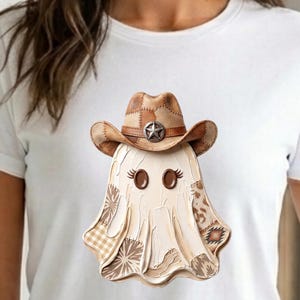 May include: A white t-shirt featuring a graphic of a ghost wearing a brown cowboy hat with a star emblem. The ghost has brown button eyes with eyelashes and is decorated with various tan and brown patterned patches including gingham and geometric prints.