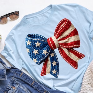 May include: Light blue t-shirt featuring a large, knitted bow design. The bow is in the colors of the American flag: red and white stripes on one side, and blue with white stars on the other. The shirt is displayed on a white surface.