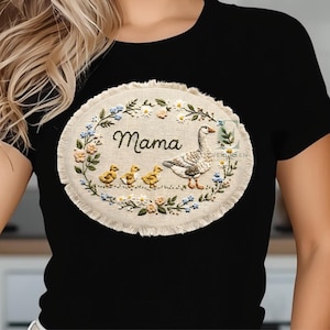 May include: Black t-shirt featuring an embroidered oval design with the word "Mama". The design includes a goose, three ducklings, and floral accents in blue, yellow, and cream colors. The design is surrounded by a fringed edge.