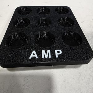 Puede incluir: Bandeja negra con nueve hendiduras circulares y las letras "AMP" en blanco. La bandeja tiene una superficie texturizada y está diseñada para guardar objetos pequeños. La bandeja está hecha de un material duradero.