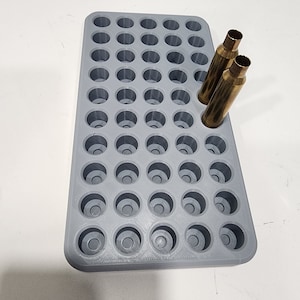 May include: A grey ammunition loading tray with numerous circular compartments. Two brass bullet casings are partially inserted. The tray is designed for organising ammunition for reloading. The background is a white surface.