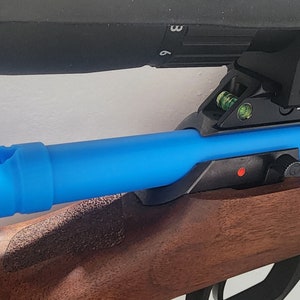 May include: A rifle with a wooden stock and a blue barrel cover. A black scope is mounted on top. The rifle has a red dot and other metal components. The blue barrel cover has a hole near the end.