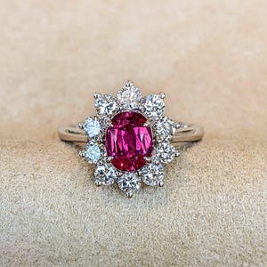 Puede incluir: Un anillo de plata con una gema central magenta de corte ovalado, rodeada por un halo de gemas redondas y transparentes más pequeñas. El anillo se presenta sobre un fondo neutro, resaltando los colores vibrantes de la joya.