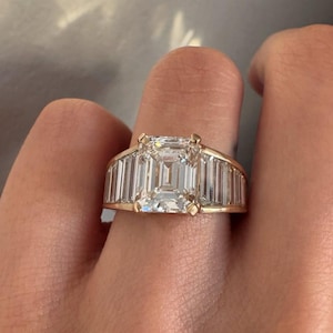May include: A gold ring featuring a large, rectangular-cut diamond center stone, flanked by smaller, rectangular diamonds. The ring's band is also gold. The ring is shown on a finger.