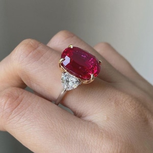 May include: A striking ring featuring a large, oval-cut ruby in a gold setting, flanked by two triangular diamonds. The ruby is a deep red hue, and the diamonds are clear. The ring is set on a silver band.