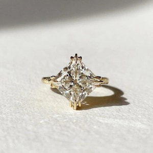 May include: A gold ring featuring a large, square-cut diamond. The diamond is clear and multifaceted, catching the light. The band has an intricate, twisted design. The ring is set against a white background, highlighting its sparkle.