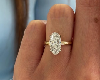 May include: A gold band ring featuring a large, oval-cut diamond. The diamond is set with four prongs and sparkles brightly. The ring is shown on a person's finger, with a light blue background.