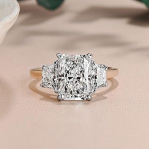 May include: A radiant cut diamond engagement ring with a gold band. The ring features a large, rectangular center stone flanked by two smaller, trapezoid-shaped diamonds. The diamonds sparkle against a soft, neutral background.