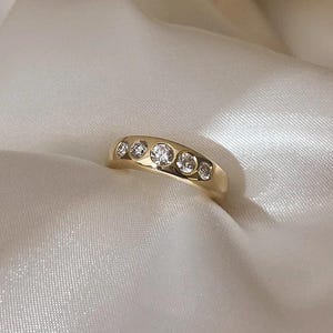 Op de afbeelding: Een gouden ring met vijf heldere, ronde edelstenen in een rij. De ring wordt gepresenteerd op een witte, zijdeachtige stof, waardoor een contrast ontstaat tussen het warme goud en de koele, gladde achtergrond. De edelstenen schitteren.