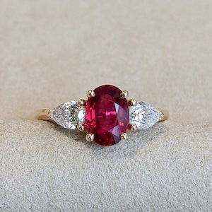 Op de afbeelding: Een gouden ring met een grote ovaal geslepen robijn als middensteen, geflankeerd door twee peervormige diamanten. De robijn heeft een dieprode kleur en de diamanten zijn helder. De ring is geplaatst tegen een neutrale achtergrond.