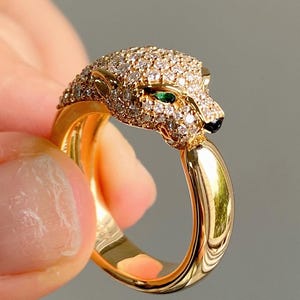 Könnte beinhalten: Ein goldfarbener Ring mit einem Pantherkopf-Design. Der Pantherkopf ist mit kleinen, funkelnden, klaren Steinen besetzt, mit einem grünen Stein für das Auge und schwarzen Akzenten. Der Ring wird von einer Hand gehalten.