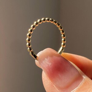 May include: A gold ring featuring a beaded design. The ring is held up by a person's finger, showcasing the ring's circular shape and the individual beads that make up the band. The ring has a polished, shiny finish.