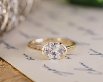 Può includere: Un anello d'oro con un grande diamante taglio ovale. Il diamante è incastonato in una montatura a castone e la fascia ha un design semplice e lucido. L'anello è esposto su una superficie color crema con una leggera scritta blu.