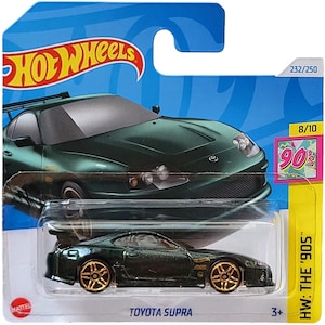 Puede incluir: Un coche de modelo fundido a presión Toyota Supra verde oscuro con ruedas doradas, empaquetado en una tarjeta Hot Wheels azul y amarilla. La tarjeta presenta el logotipo de Hot Wheels, el texto "Toyota Supra" y "HW: THE '90s".