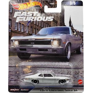 May include: A silver 1970 Chevrolet Nova SS diecast car model from the Fast & Furious franchise. The car is displayed against a black and white brick background. The car has a black stripe across the hood and the words "Fast & Furious" are printed on the card.