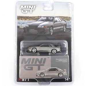 Puede incluir: Un coche modelo en miniatura plateado Nissan Skyline GT-R (R32) en una vitrina de plástico transparente. El embalaje presenta el logotipo "MINI GT" y el texto "Nissan Skyline GT-R (R32) VeSide Combat C-1 Combat Grey". El modelo es para mayores de 14 años.