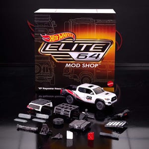 May include: A white and black Hot Wheels '17 Toyota Tacoma model with a red stripe, displayed with various mod shop parts. The box behind the truck reads "ELITE 64 MOD SHOP". Includes a small white and red fuel container.