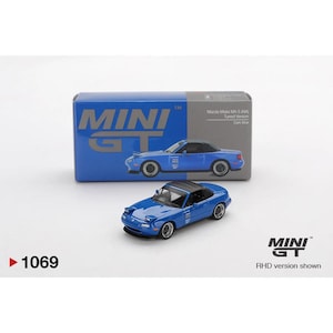 May include: A blue diecast model car with a black roof, displayed in front of its box. The box is blue with the words "MINI GT" in white. The model car is a Mazda Miata MX-5, and the box indicates it is the "Tuned Version".