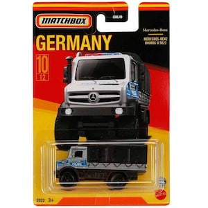 Puede incluir: Un modelo de coche de juguete de fundición a presión Matchbox a escala 1:64 de un Mercedes-Benz Unimog U 5023 blanco con una caja negra y marcas de policía azules y blancas. El coche está en una tarjeta amarilla y roja con las palabras "Alemania" y "Matchbox" impresas en ella.