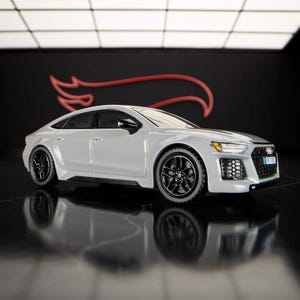 May include: A white Audi RS 7 Sportback diecast model car with black wheels. The car is on a black surface with a red Hot Wheels logo in the background.