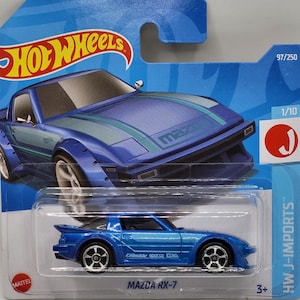 Könnte beinhalten: Ein blaues, aus Metall gegossenes Spielzeugautomodell eines Mazda RX-7 mit einem grün-weißen Rennstreifen. Das Auto befindet sich auf einem blauen Hintergrund mit den Worten "Hot Wheels" und "HW J-Imports", die auf der Verpackung aufgedruckt sind. Das Auto ist ein Modell im Maßstab 1:10.