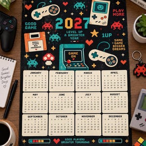 Puede incluir: Un calendario de pared para 2027 con fondo negro y gráficos coloridos de arte de píxeles que muestran mandos de videojuegos, una consola portátil, una máquina recreativa y un disquete. El texto incluye "GOOD GAME", "LEVEL UP A BRIGHTER YEAR", "1UP" y "GOOD PLAYERS BRIGHTER TOMORROWS".