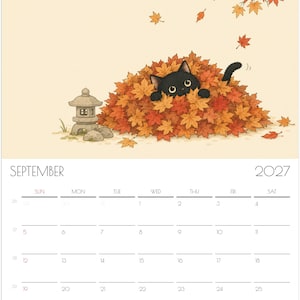 Puede incluir: Un calendario de pared para septiembre de 2027 con una ilustraci&oacute;n de un gato negro asom&aacute;ndose desde un gran mont&oacute;n de hojas de arce oto&ntilde;ales naranjas y rojas, con una peque&ntilde;a linterna de piedra cerca y hojas cayendo sobre un fondo crema.