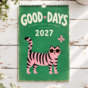 May include: A 2027 wall calendar with a green background and a spiral binding at the top. The cover features the text "GOOD DAYS A HAPPY LITTLE CALENDAR" in white, a pink cat with black stripes wearing round sunglasses, and a small pink butterfly.