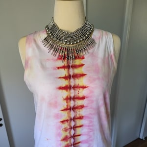 May include: White sleeveless shirt with a tie-dye pattern in shades of pink, red, and yellow. The shirt is decorated with a silver beaded necklace.