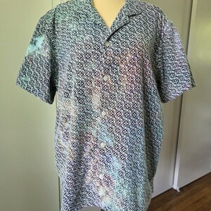 May include: A short-sleeved button-down shirt with a geometric pattern in shades of blue, white, and purple. The shirt is displayed on a mannequin. The collar is open, and the shirt has white buttons.