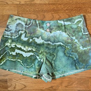 May include: High-waisted corduroy shorts with a unique watercolor-like pattern in shades of green, blue, and white. The shorts feature two front pockets, two silver buttons, and a small folded hem. The fabric has a ribbed texture.