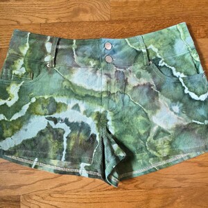 May include: Green and blue tie-dye denim shorts with a light blue wash. The shorts have a button and zipper closure and two front pockets.